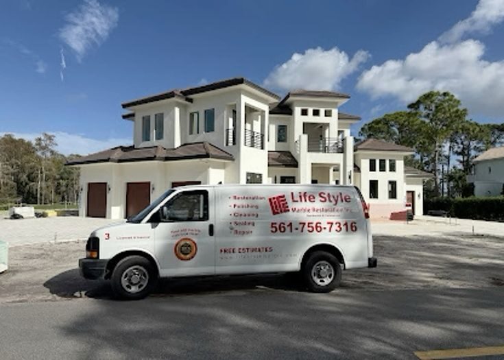 A white Lifestyle Marble Restoration van is parked in front of a modern, light-colored two-story house or building. The van has the "Lifestyle Marble Restoration" logo and text "Floors, Countertops, Showers" and a phone number (561-756-7316)(though it's a bit blurry). The setting appears to be a sunny day.